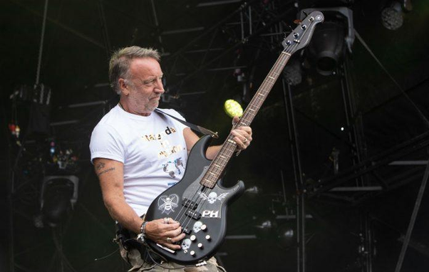 Peter Hook con pelo canoso y barba corta toca un bajo eléctrico negro en un escenario. Viste una camiseta blanca y pantalones de camuflaje. El bajo está distintivamente decorado con calaveras blancas, abejas y las letras 'PH'; un limón o lima verde brillante cuelga de su clavijero. El hombre tiene una expresión concentrada mientras toca, con un tatuaje visible en su brazo derecho y varios anillos. El fondo es oscuro, con estructuras metálicas y focos de escenario apenas visibles.