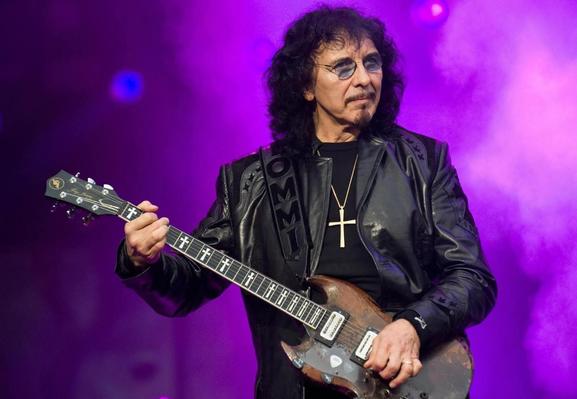 Tommy Iommi toca la guitarra. Tiene el cabello largo, viste una chaqueta de cuero negra y se observa un colgante con una cruz en su cuello.