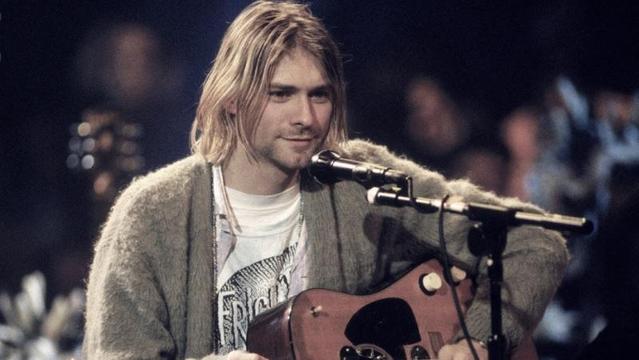 Kurt Cobain sentado frente a un micrófono con una guitarra acústica.