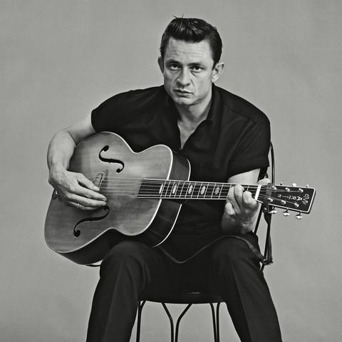 Foto en blanco y negro de Johnny Cash, quien está sentado, luce cabello corto muy bien peinado mientras toca una guitarra acústica que sostiene en sus piernas.