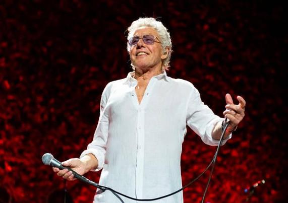Roger Daltrey de pie sonríe, luce cabello blanco y una camisa blanca con un micrófono en la mano.