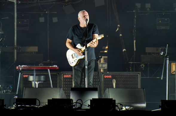 David Gilmour sobre un escenario canta frente al micrófono mientras toca la guitarra.