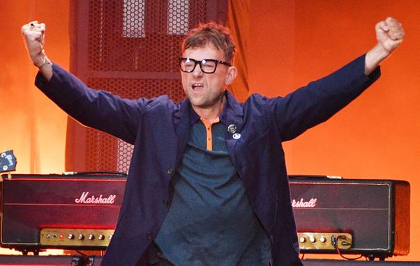 Damon Albarn, vocalista de Blur, foto desde la cintura hacia arriba, está con las mano empuñadas y los brazos extendidos en señal de celebración. Luce lentes anchos y el cabello desornado. Viste una chaque azul oscuro.