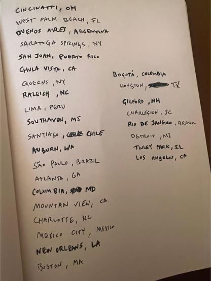 "Una página de cuaderno o papel blanco con una lista de ciudades y sus respectivos estados o países, escrita a mano con tinta negra. La lista está organizada en dos columnas principales, con la segunda columna comenzando más abajo en la página. Entre las ciudades mencionadas se encuentran Cincinnati, OH; Buenos Aires, Argentina; San Juan, Puerto Rico; Lima, Perú; São Paulo, Brasil; Ciudad de México, México; Bogotá, Colombia; Houston, TX (con un pequeño garabato sobre parte del nombre); Río de J…