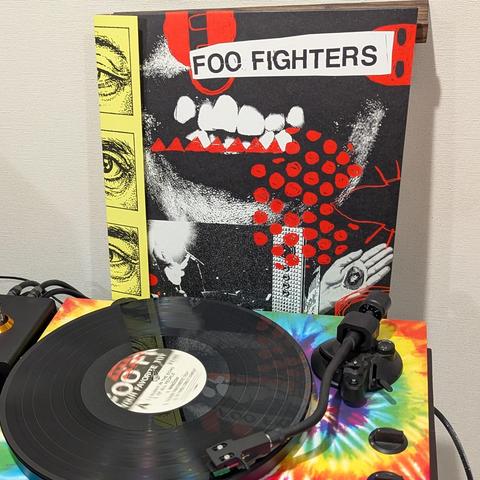 Una imagen muestra la portada de un álbum de Foo Fighters de pie, detrás de un tocadiscos con un disco de vinilo negro reproduciéndose.

La portada del álbum es predominantemente negra, con el nombre "FOO FIGHTERS" en una banda blanca con borde rojo en la parte superior. Presenta una ilustración estilizada y agresiva con grandes dientes blancos y encías rojas en el centro. A la izquierda, hay una franja vertical amarilla con múltiples ilustraciones de ojos. A la derecha, se ven puntos rojos dis…