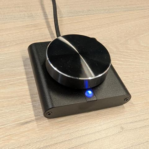 Un dispositivo electrónico rectangular de color negro mate, que parece ser un controlador de audio o DAC, descansa sobre una mesa de madera clara con vetas visibles. En su parte superior central, destaca una gran perilla circular negra con un borde plateado brillante y una superficie superior con un acabado metálico cepillado que refleja la luz. Debajo de la perilla, en el frente del dispositivo, se ilumina un pequeño LED azul brillante. Un cable negro trenzado sale de la parte trasera del disp…