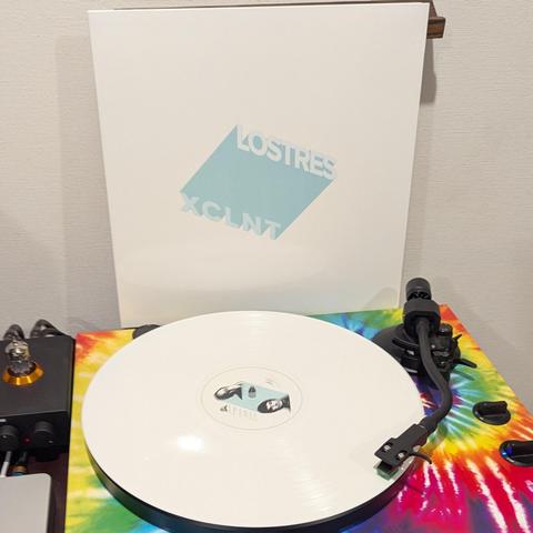 "La imagen muestra un tocadiscos en primer plano con un vinilo blanco girando. La etiqueta central del vinilo tiene un diseño abstracto en tonos claros, con lo que parecen ser dos rostros o figuras. La base del tocadiscos es muy llamativa, con un vibrante patrón tie-dye que incluye colores brillantes como rojo, amarillo, azul, verde y morado. Detrás del tocadiscos, se apoya verticalmente una portada de álbum mayormente blanca. En el centro de la portada, hay una forma rectangular azul clara con…