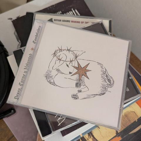 Aquí tienes una descripción de texto alternativo para la imagen:

Una caja de CD transparente descansa sobre una pila de otros álbumes. La carátula del CD es blanca y presenta un dibujo lineal de un cordero acostado. El cordero lleva una corona de espinas en la cabeza y tiene una estrella dorada de múltiples puntas en su costado. El dibujo tiene un estilo boceto, con líneas negras y la estrella con color. En el lomo del CD se lee "Deseo, Carne y Voluntad" y "Candelabro", junto a una pequeña est…