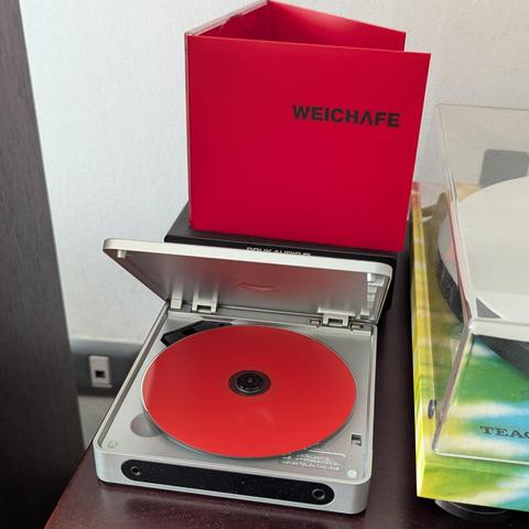 Una imagen muestra un reproductor de CD portátil de color gris plateado sobre una superficie de madera oscura. La tapa del reproductor está abierta, revelando un disco de color rojo brillante en su interior. Encima del reproductor, sobre una base negra, hay una caja o folleto de CD de color rojo intenso con la palabra "WEICHAFE" escrita en negro. A la derecha, se observa parcialmente otro equipo de audio con una cubierta transparente y detalles en verde y amarillo, con la marca "TEAC" visible. …