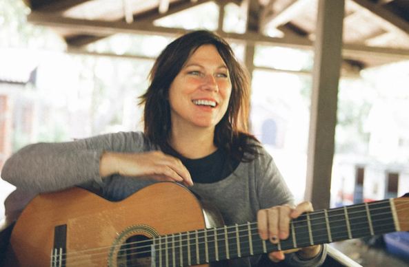Kim Deal sonriente con cabello oscuro y ojos verdes sostiene una guitarra clásica de madera clara. Su rostro irradia alegría con una amplia sonrisa que muestra los dientes, y mira ligeramente hacia la derecha. Sus manos están posicionadas sobre el instrumento como si estuviera tocando. El fondo está suavemente desenfocado, revelando una estructura de madera y vegetación, sugiriendo un entorno exterior o semi-abierto.