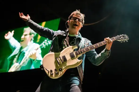 Rivers Cuomo enérgico, con gafas y una chaqueta vaquera, toca una guitarra eléctrica color crema en un escenario. Canta con la boca abierta, su brazo izquierdo levantado y la palma de la mano abierta. La guitarra es de doble cutaway, con pastillas oscuras y herrajes cromados. Detrás de él, una gran pantalla proyecta una imagen borrosa en tonos verdes de un hombre con una guitarra, también con los brazos levantados, replicando la energía del momento. El fondo es oscuro, con un foco principal sob…