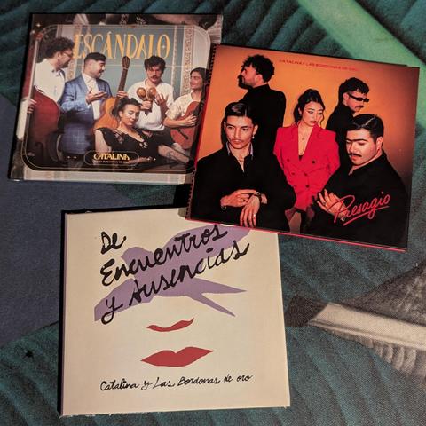La imagen muestra tres portadas de álbumes de música del grupo "Catalina y Las Bordonas de Oro".

1. **Portada superior izquierda, "Escándalo":** Presenta a seis músicos (cinco hombres y una mujer) en un estilo retro, posando con instrumentos como un contrabajo, guitarras y maracas. Visten ropa elegante y la escena tiene un aire festivo y clásico.
2. **Portada superior derecha, "Presagio":** Muestra a cinco miembros del grupo (cuatro hombres y una mujer) posando de forma moderna y sofisticada c…