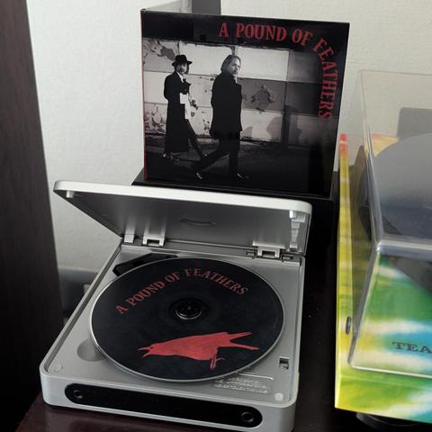 Una imagen muestra un reproductor de CD compacto de color plateado con su tapa abierta. Dentro hay un CD negro con el título "A POUND OF FEATHERS" en letras rojas y la silueta de un pájaro rojo. Detrás del reproductor, se ve la carátula del álbum, que presenta una foto en blanco y negro de dos hombres caminando, uno con sombrero, y el mismo título en letras rojas. A la derecha, se observa una parte de otra caja de álbum con una cubierta transparente y un patrón de colores vibrantes.