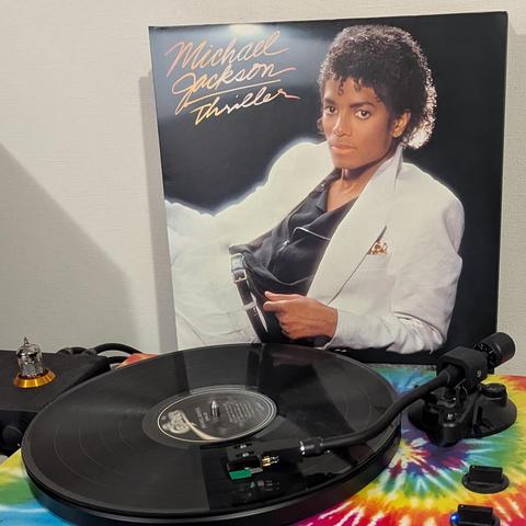 La imagen muestra la portada del álbum 'Thriller' de Michael Jackson, de pie, detrás de un tocadiscos. La portada presenta a Michael Jackson sentado, con una chaqueta de traje blanca y camisa negra. Con su característico cabello rizado, mira directamente al espectador. Sobre un fondo negro, el título 'Michael Jackson Thriller' está escrito en cursivas doradas en la esquina superior izquierda.

En primer plano, un tocadiscos reproduce un disco de vinilo negro. El brazo del tocadiscos, con una ag…