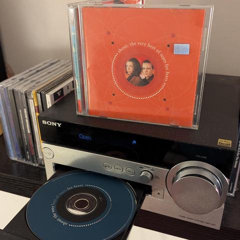 Una vista de cerca de un sistema de audio doméstico Sony plateado y negro con la bandeja de CD abierta. Un CD azul oscuro con el título "Shout: The Very Best of Tears for Fears" está parcialmente fuera de la bandeja. Encima del reproductor, se encuentra la caja de jewel case del mismo álbum, con una carátula naranja vibrante. La carátula muestra un círculo central con los retratos de dos hombres (Curt Smith y Roland Orzabal) y el título del álbum en un arco alrededor de ellos, con un diseño de …