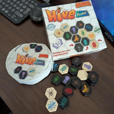 La imagen muestra el juego de mesa "Hive Pocket Ultimate" desplegado sobre una mesa de madera. En la parte superior derecha se encuentra la caja del juego, blanca con el logo "Hive" grande en naranja y "Pocket" en azul, rodeado de un patrón hexagonal y ejemplos de las fichas de insectos. La caja indica que incluye las tres expansiones y una bolsa de viaje. A la izquierda, hay una pequeña bolsa de tela blanca, redonda, con el mismo logo y algunas fichas de insectos impresas. En la parte inferior…
