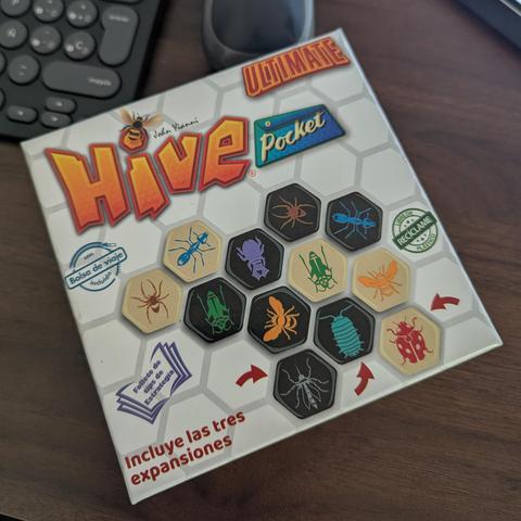 Una caja del juego de mesa "Hive Pocket Ultimate" se muestra ligeramente inclinada sobre un escritorio de madera oscura. La caja es predominantemente blanca con un patrón hexagonal de fondo. El logo principal "Hive" es grande, naranja y tiene textura de panal, con la palabra "Ultimate" en rojo arriba y "Pocket" en azul a la derecha. Una abeja vuela cerca del logo. La caja exhibe varias fichas hexagonales de juego, algunas de color crema y otras negras, cada una con una ilustración de un insecto…