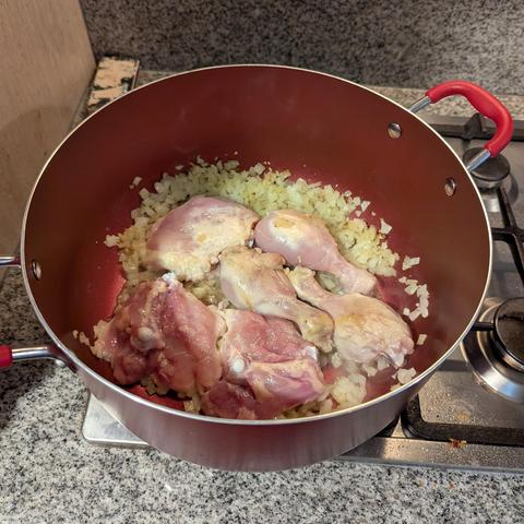 Una olla o sartén profunda de color rojo intenso con asas rojas está sobre una estufa de gas. En su interior, varias piezas de pollo crudo o ligeramente cocido (muslos y piernas) se mezclan con abundante cebolla blanca picada y translúcida, sugiriendo que se están sofriendo. La olla se apoya sobre una encimera de cocina de granito gris moteado.