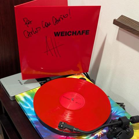 Un disco de vinilo color rojo reproduciéndose en una tornesa colorida. En la parte de atrás la carátula del vinilo, completamente roja, en letras negras divw "WEICHAFE". ESTÁ rayada con lápiz negro en letraanuscrita dice "Con cariño para Carlos" y tiene la firma de Angelo Pierattini.