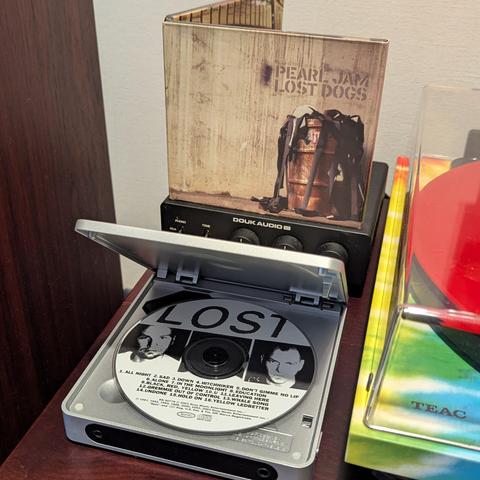 Una vista cercana de un reproductor de CD portátil plateado con la tapa abierta, revelando un CD de Pearl Jam "Lost Dogs". El CD tiene la palabra "LOST" en letras grandes y dos retratos en blanco y negro de miembros de la banda. Detrás del reproductor, se ve la carátula del álbum "Pearl Jam Lost Dogs", que muestra un barril oxidado con correas sobre un fondo que parece madera envejecida. Debajo de la carátula hay un pequeño amplificador de audio negro de la marca Douk Audio con varios botones y…