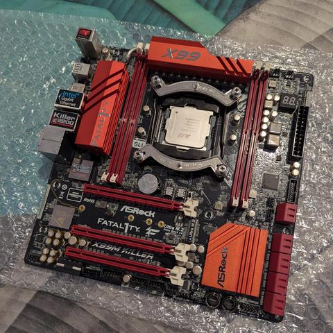 Una placa base (motherboard) ASRock Fatal1ty X99M Killer de color negro con detalles prominentes en rojo, reposando sobre plástico de burbujas. En el centro, un procesador Intel plateado está visible en su zócalo, rodeado por un soporte metálico. A ambos lados del procesador, se aprecian ocho ranuras DIMM rojas para memoria RAM. Varios disipadores de calor rojos están distribuidos por la placa: uno grande en la parte superior izquierda con la palabra "FATALITY", otro en la superior derecha que …