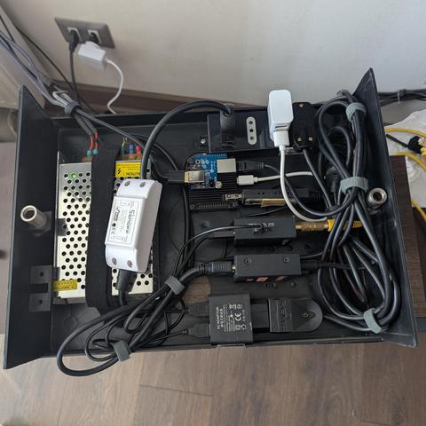 La imagen muestra el interior de una caja rectangular oscura, posiblemente un gabinete, lleno de varios componentes electrónicos y cables. En el lado izquierdo, hay una fuente de alimentación metálica perforada y plateada, con cables conectados y una etiqueta de advertencia. Junto a ella, un dispositivo blanco con la marca 'Sonoff' está conectado, probablemente un interruptor inteligente o relé. En la parte superior central, se observa una pequeña placa base de circuito azul con puertos (Ethern…