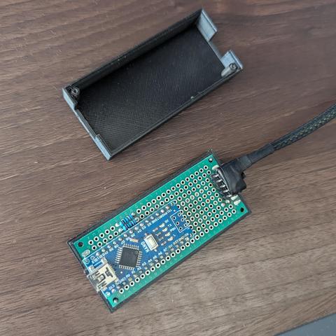 Una placa de circuito verde, que contiene un módulo Arduino Nano azul con un puerto USB mini-B, está conectada a un cable trenzado negro. La placa principal tiene una zona de protoboard con múltiples orificios. A su lado, hay una carcasa negra abierta, impresa en 3D, que parece diseñada para alojar el circuito. Todo descansa sobre una superficie de madera de tono medio.