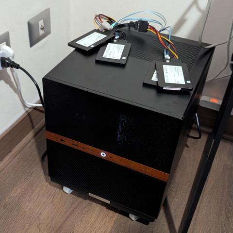 Una caja de ordenador compacta y negra, con una franja horizontal de madera o imitación madera y un botón de encendido circular iluminado en blanco en el centro de su panel frontal. Sobre la parte superior de la caja, hay cuatro unidades de estado sólido (SSD) negras, conectadas con un manojo de cables SATA de colores que se introducen en la parte trasera de la caja. La caja descansa sobre pequeñas ruedas o patas blancas en un suelo de madera clara. En la pared de fondo, visible a la izquierda,…