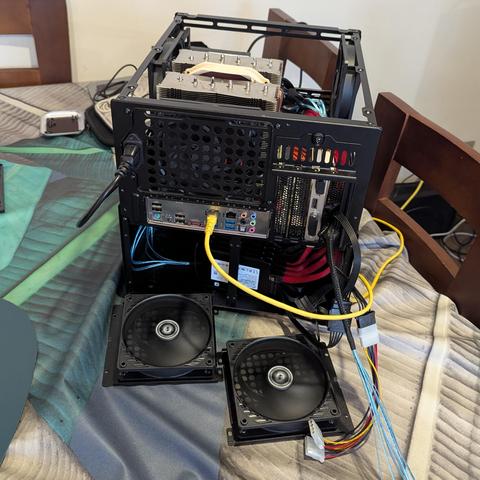 Una torre de computadora (PC) de color negro, abierta y en proceso de ensamblaje o mantenimiento, reposa sobre una superficie acolchada con un patrón. La parte trasera de la torre es visible, mostrando la placa madre con sus puertos (Ethernet, USB, audio) y una tarjeta de expansión instalada. Un cable de alimentación negro está conectado a la fuente de poder y un cable Ethernet amarillo a la placa madre. En el interior, se destaca un voluminoso disipador de CPU de doble torre con ventiladores d…