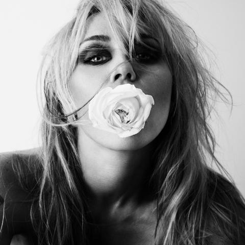 Primer plano en blanco y negro de Miley Cyrus con el cabello rubio, largo y ligeramente despeinado, que enmarca su rostro. Tiene una rosa blanca entre los labios, que mira directamente al frente con una expresión intensa. Sus ojos están maquillados con un estilo ahumado oscuro. Se aprecian sus hombros desnudos.