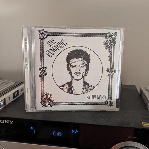 Una caja de CD de música está apoyada sobre un reproductor de audio oscuro. La carátula del CD es un dibujo en blanco y negro de Bruno Mars, con pelo rizado, una bandana blanca en la frente y bigote. Su imagen está dentro de un círculo, rodeado por un marco decorado con cadenas y rosas en las esquinas. Arriba del círculo se lee "THE ROMANTIC" en letras estilizadas, y debajo "BRUNO MARS". En el lomo del CD también se lee "THE ROMANTIC: BRUNO MARS". En el reproductor de audio, se ve parte de la m…