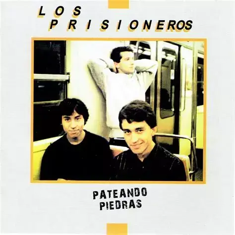 Portada del álbum 'Pateando Piedras' de Los Prisioneros. Sobre un fondo blanco, una fotografía rectangular enmarcada en amarillo muestra a los tres jóvenes integrantes de la banda posando de manera informal dentro de un vagón de metro o tren. En el fondo, uno de ellos, con una camisa clara, está de pie o apoyado, con los brazos detrás de la cabeza. En primer plano, los otros dos están sentados o agachados; el de la izquierda tiene el pelo oscuro, viste una camisa negra y mira con expresión neut…