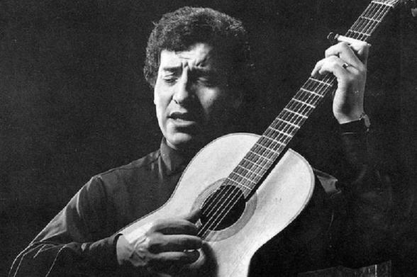 Una fotografía en blanco y negro muestra a Víctor Jara en plena interpretación musical. Tiene el pelo oscuro y rizado, y su rostro muestra una expresión de profunda concentración o emoción, con los ojos cerrados y la boca ligeramente abierta. Viste una camisa oscura de cuello alto. Sostiene una guitarra clásica de cuerpo claro. Su mano izquierda presiona las cuerdas en el mástil, donde se aprecia un capo y un anillo en su dedo anular. Su mano derecha rasguea las cuerdas cerca de la boca de la g…