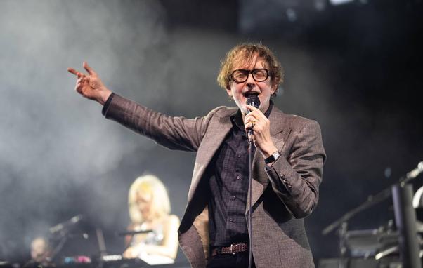 Jarvis Cocker canta en un escenario, con el pelo castaño claro, rizado y algo despeinado, y gafas de montura gruesa y oscura. Viste una chaqueta de traje de color gris parduzco o tweed, sobre una camisa oscura a cuadros. Sostiene un micrófono negro con su mano derecha cerca de la boca, mientras su mano izquierda está extendida hacia la izquierda, formando un signo de 'V' con los dedos índice y medio levantados. Su boca está abierta y sus ojos cerrados o entrecerrados, sugiriendo que está cantan…