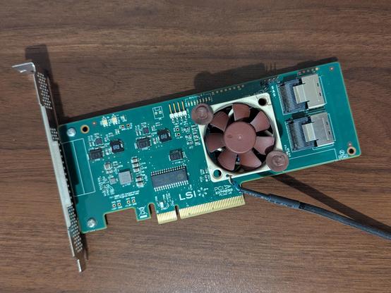 Una tarjeta de expansión de computadora (PCIe) de color verde oscuro, rectangular, orientada horizontalmente sobre una superficie de madera. En su lado izquierdo tiene un soporte metálico plateado perforado. En el centro de la tarjeta hay un pequeño ventilador de color marrón o bronce con seis aspas. Hacia el lado derecho superior, hay dos puertos de conexión SAS (Serial Attached SCSI) apilados verticalmente, de color plateado. La tarjeta está cubierta de varios componentes electrónicos pequeño…