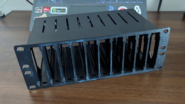 Un soporte o bahía de almacenamiento de color negro, fabricado en plástico con una ligera textura, diseñado para montaje en rack. Presenta una fila de múltiples ranuras verticales para organizar discos duros (HDD/SSD) o componentes electrónicos. La parte superior tiene ranuras horizontales para ventilación. En los laterales, hay pestañas con agujeros ovalados para su fijación. El objeto descansa sobre una superficie de madera, con el teclado desenfocado de un portátil visible al fondo.