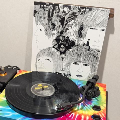 La imagen muestra un tocadiscos con un vinilo negro girando en primer plano. El tocadiscos tiene una base vibrante con un patrón tie-dye de colores rojo, amarillo, verde y azul. En el centro del disco se puede leer "PARLOPHONE". Detrás del tocadiscos, apoyada contra una pared clara, se encuentra la icónica portada del álbum "Revolver" de The Beatles. La portada es en blanco y negro, con un estilo artístico que combina dibujos lineales detallados de los rostros de los miembros de la banda con pe…