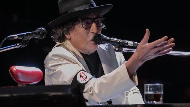 Charly García con un sombrero fedora negro y gafas de montura oscura, canta o habla intensamente en un micrófono en un escenario oscuro. Viste un blazer color crema sobre una camisa oscura. En su brazo izquierdo, lleva un brazalete negro con un círculo blanco que muestra un símbolo rojo similar a la anarquía. Su mano derecha está levantada y abierta, gesticulando. Frente a él, sobre una superficie oscura (posiblemente un piano), se ven un cojín rojo acolchado y un vaso con una bebida oscura.